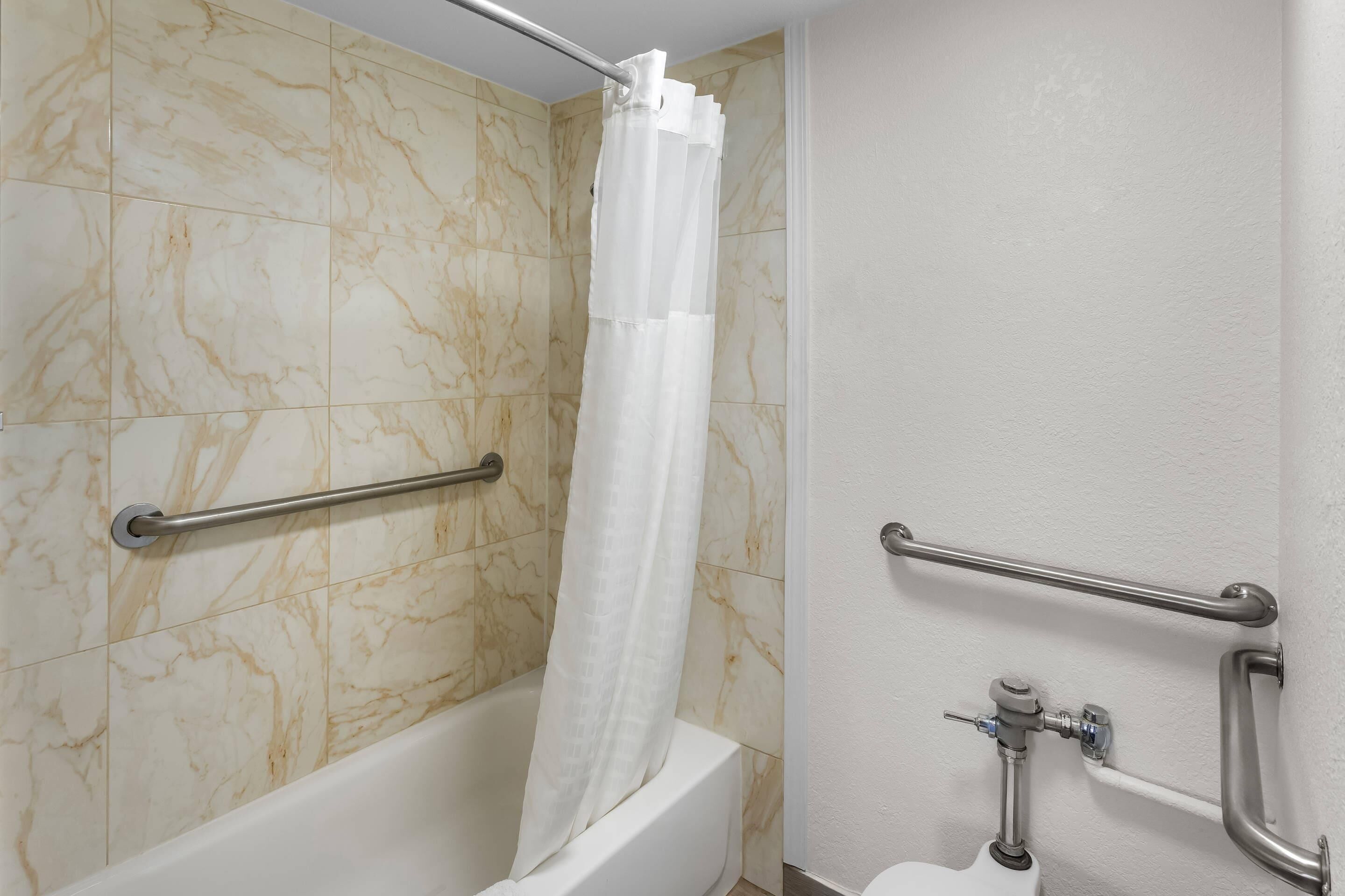 A bathroom with a bathtub, marble tiles, a white shower curtain, and grab bars.