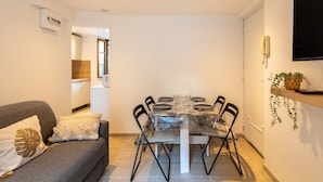 Un comedor con una mesa preparada para cuatro personas, rodeada de sillas, un sofá con cojines, un televisor montado y una planta en la pared.