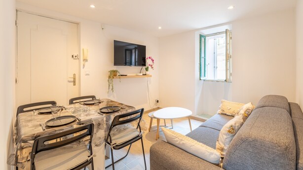 Una sala moderna con un sofá gris, una mesa de comedor con sillas, un televisor de pantalla plana montado en la pared y un ventanal con vistas.