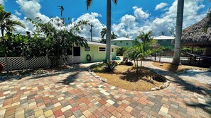 A paved courtyard with palm trees, a small building, and a thatched-roof structure.