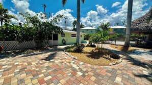 A paved courtyard with palm trees, a small building, and a thatched-roof structure. - Turtle Nest Beach Studio - Quick Walk to Beach (Fort Myers Beach)
