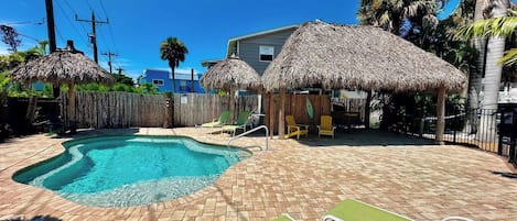 A swimming pool with a brick patio, lounge chairs, and thatched umbrellas.