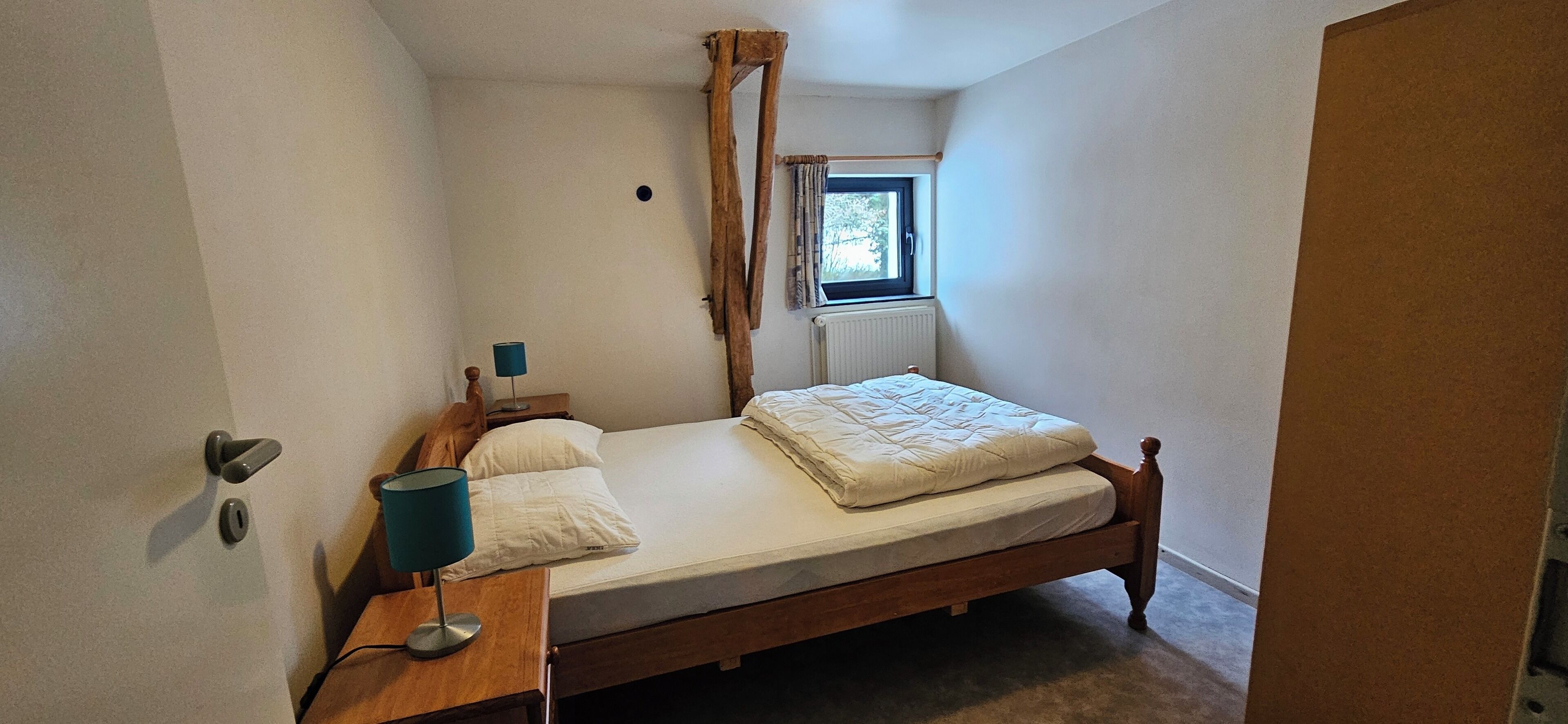 Ein kleines Zimmer mit einem Einzelbett, einem hölzernen Bettgestell, einem Nachttisch mit Lampe, einem Fenster mit Vorhang und einer Heizung.