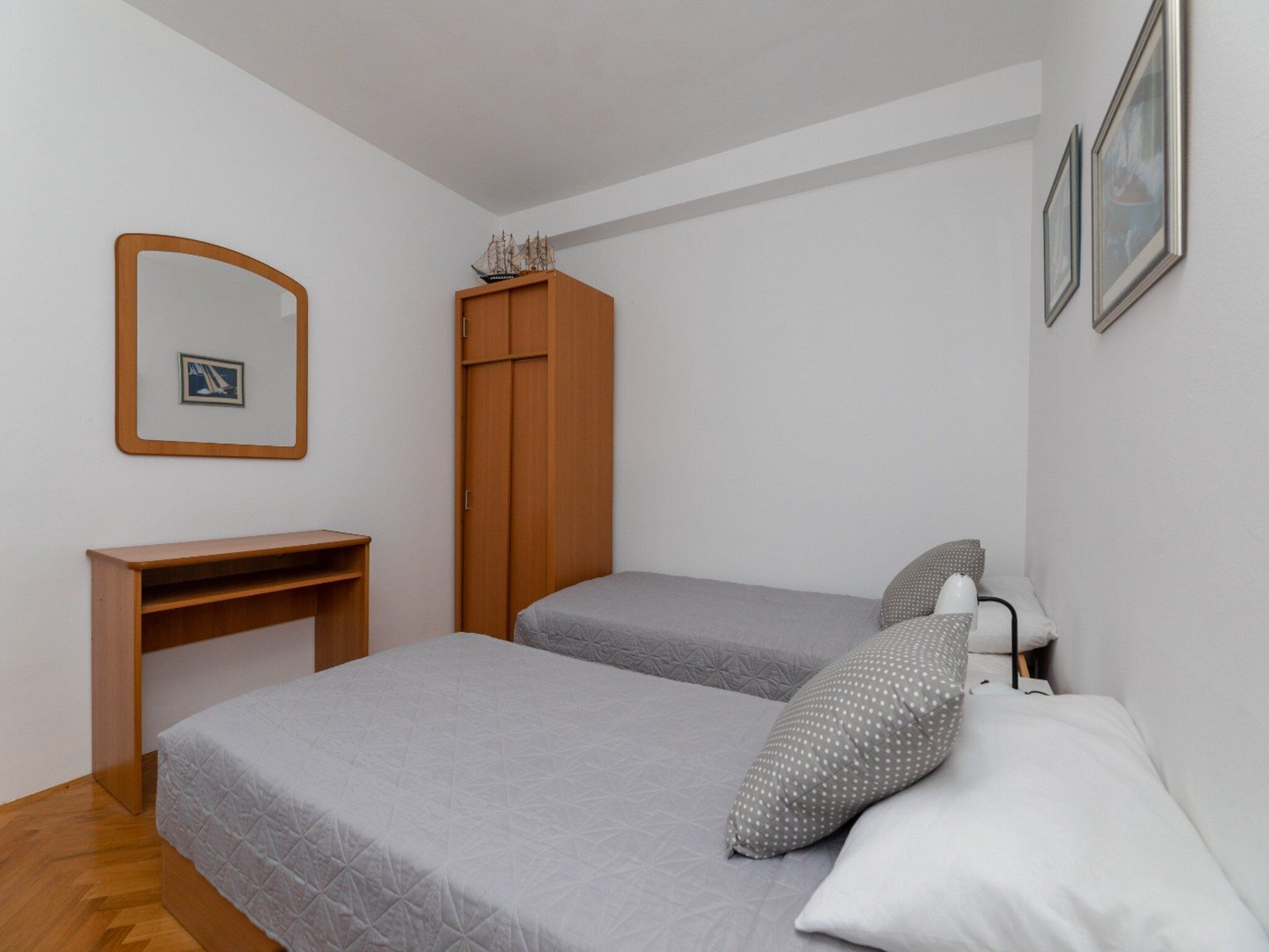 A small, neatly arranged bedroom with two beds, a wooden wardrobe, a desk, and a mirror.