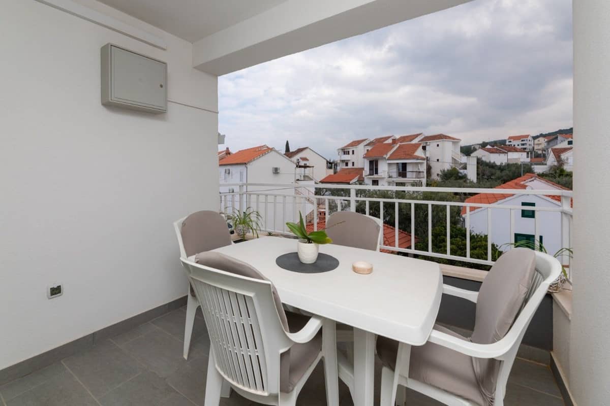A balcony with a white table and chairs, a potted plant, and a view of houses in the background.