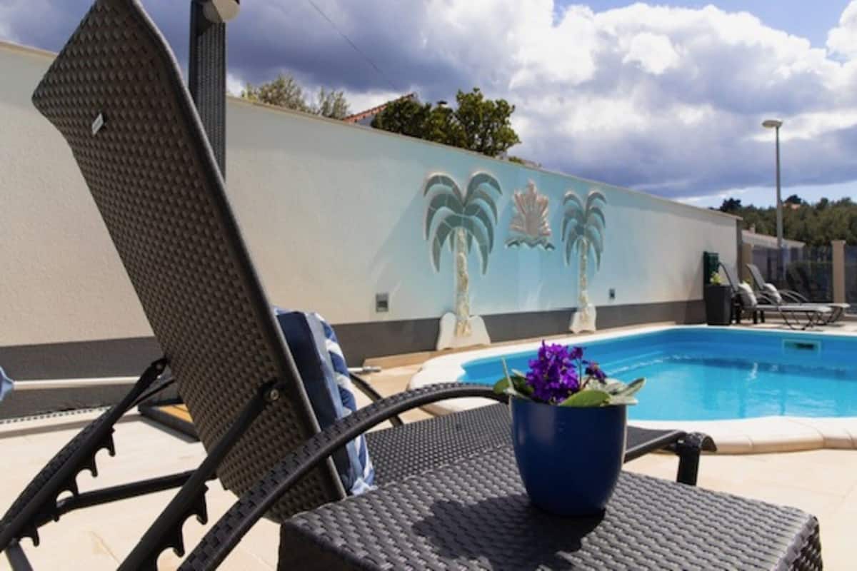 A poolside area with lounge chairs, a decorative wall with palm tree motifs, and a flower pot.