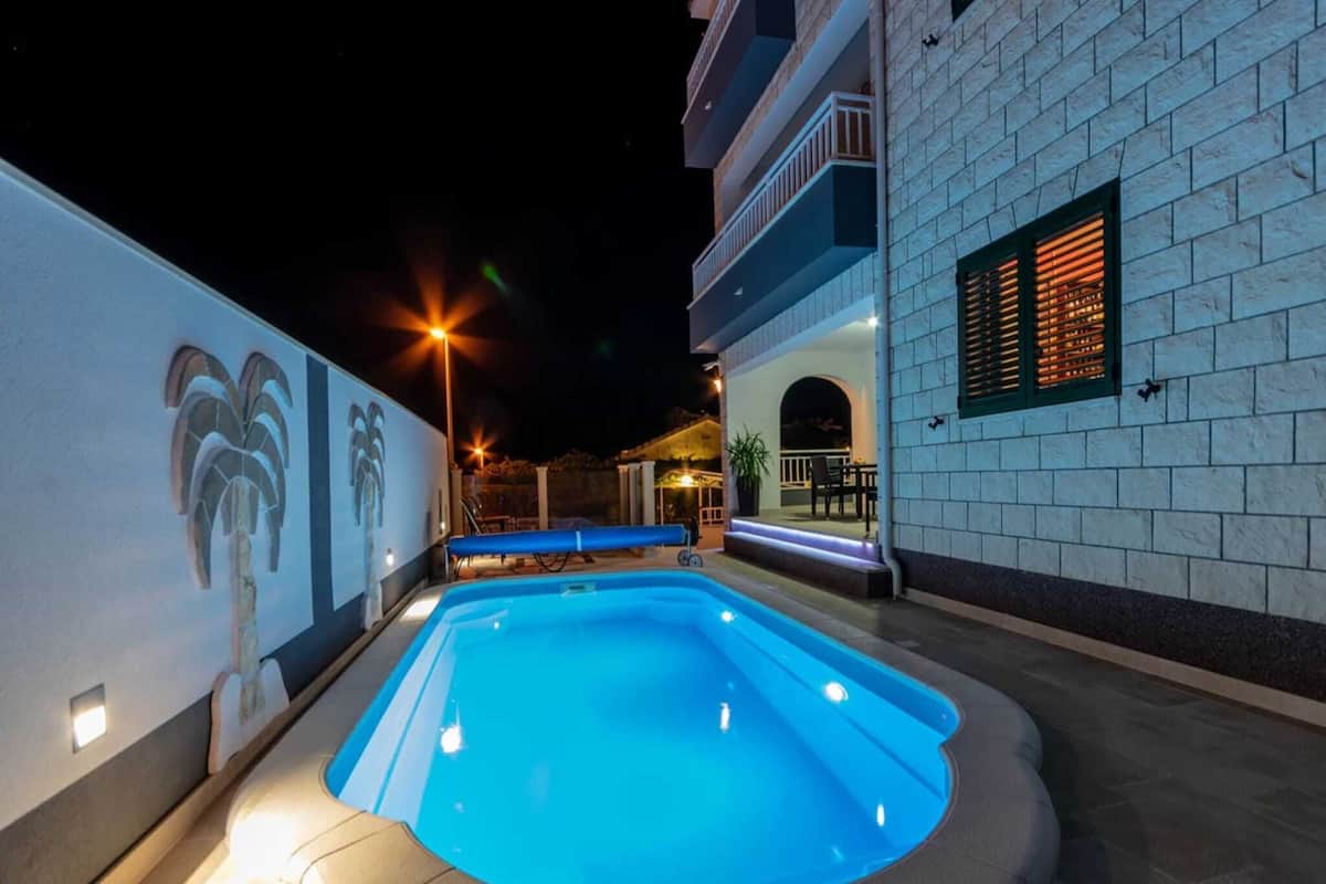 A swimming pool with a blue hue, surrounded by a white wall with a palm tree mural, and a building with a balcony and windows.