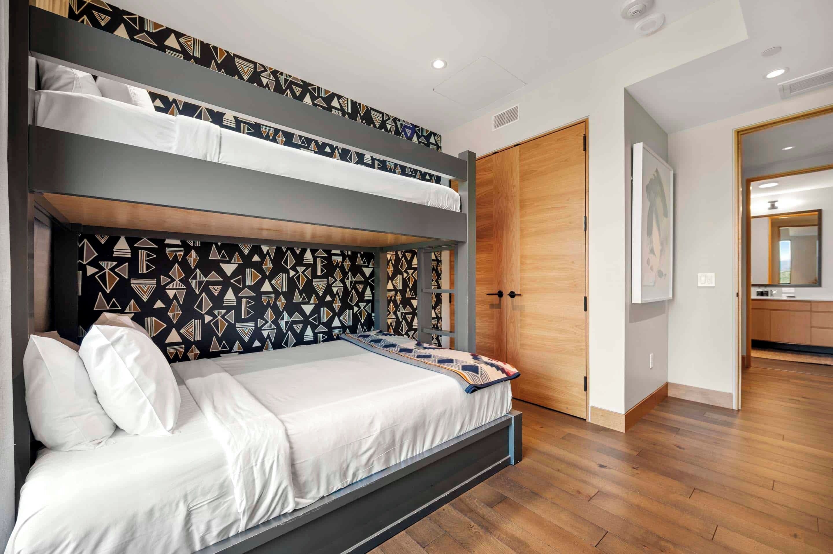 A modern bedroom with a bunk bed, patterned headboard, wooden flooring, and a door leading to a bathroom.
