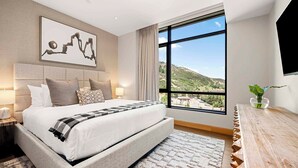 A bedroom with a large bed, a view of the outdoors, a mounted TV, and a bedside table with a plant. - One Snowmass Residence Club 403 (Snowmass)