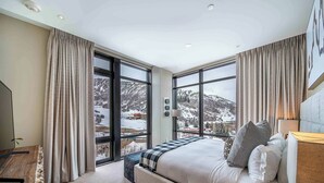 A modern hotel room with a large bed, a desk, and a view of snowy mountains through large windows.
