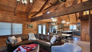 A spacious living area with a leather sectional sofa, a gray armchair, and a dining table with chairs. A kitchen with a large island and modern appliances is visible in the background.