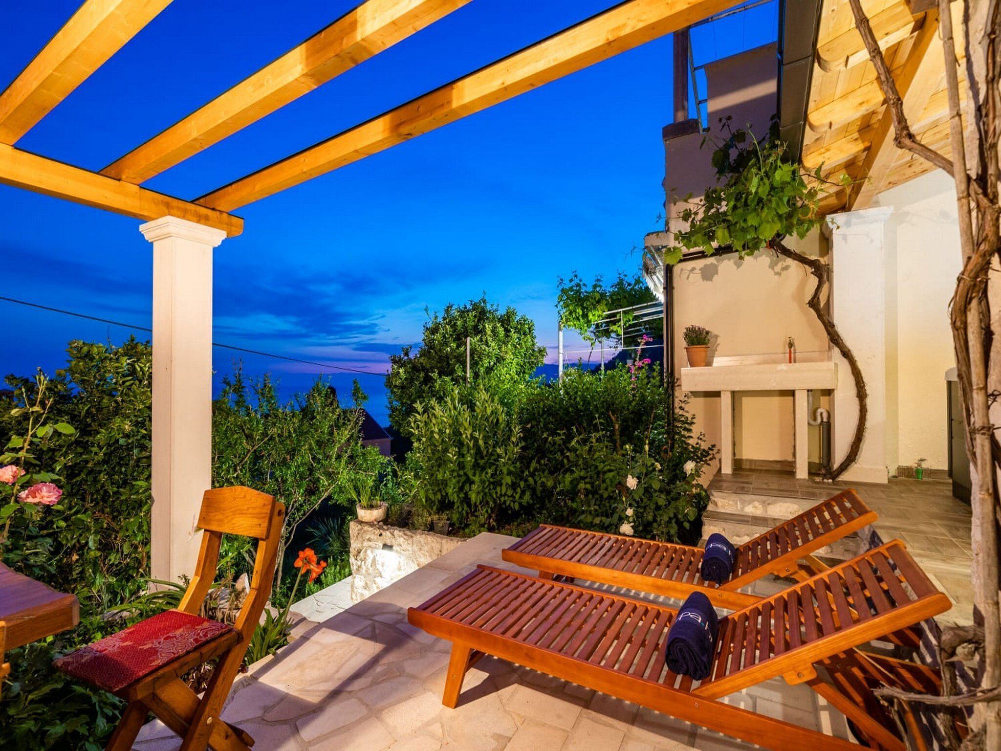 A wooden pergola with two lounge chairs and a table, surrounded by greenery and a view of the sea.