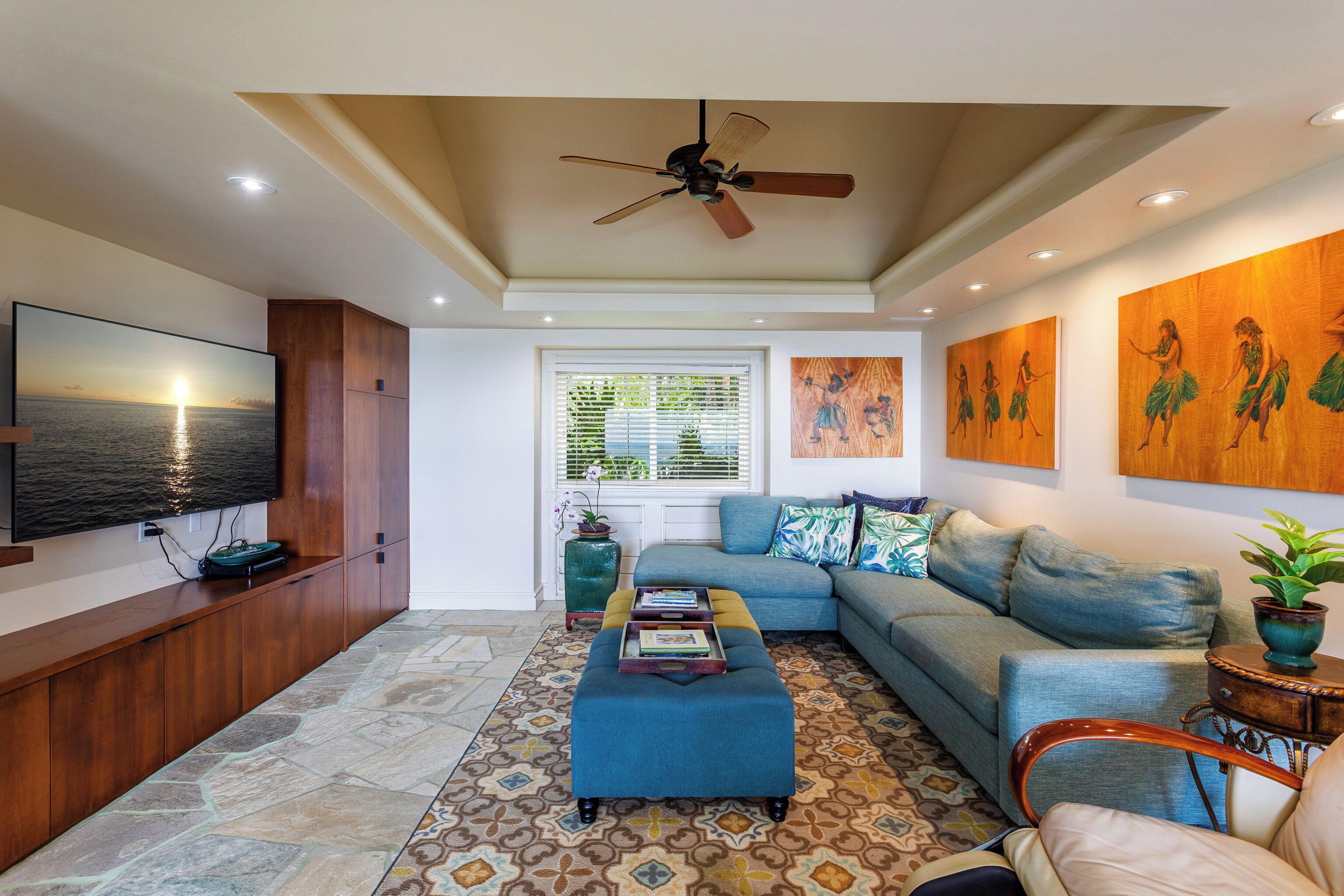 A living room with a sofa, ottoman, TV, ceiling fan, artwork, and a window with blinds.