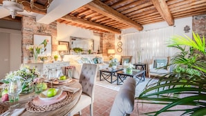 A rustic dining area with a wooden table set for a meal, surrounded by comfortable seating and a brick wall. - Valle del Sole Winery Farmhouse (Lucca)