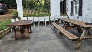 En udendørs terrasse med træborde og hvide stole, omgivet af en bygning med hvide vægge og sorte skodder.