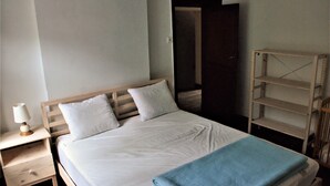 Et soveværelse med en træseng, hvide sengetæppe, et natbord med en lampe og et blåt tæppe.