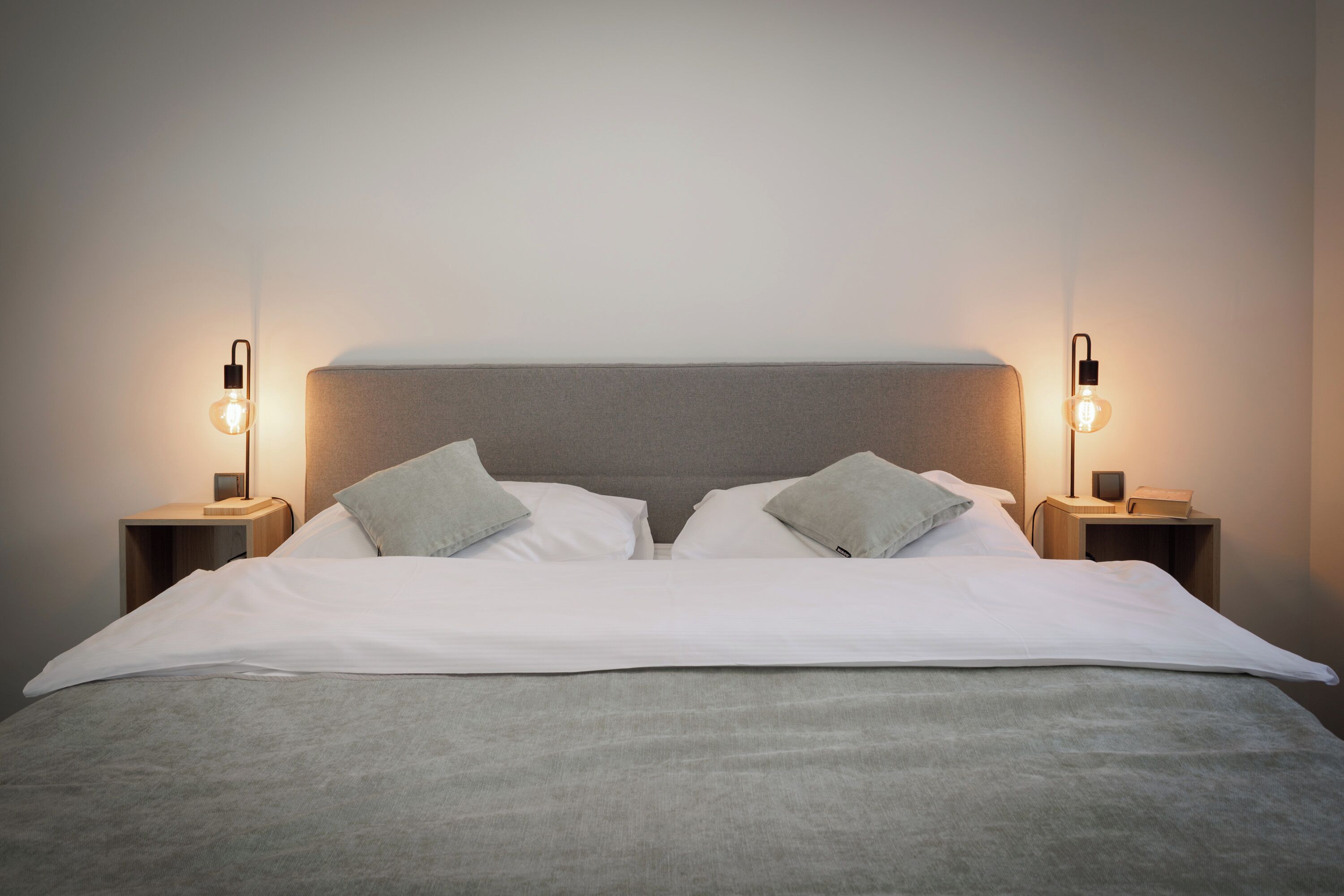 A neatly made bed with a grey headboard and two bedside tables with wall-mounted lamps.