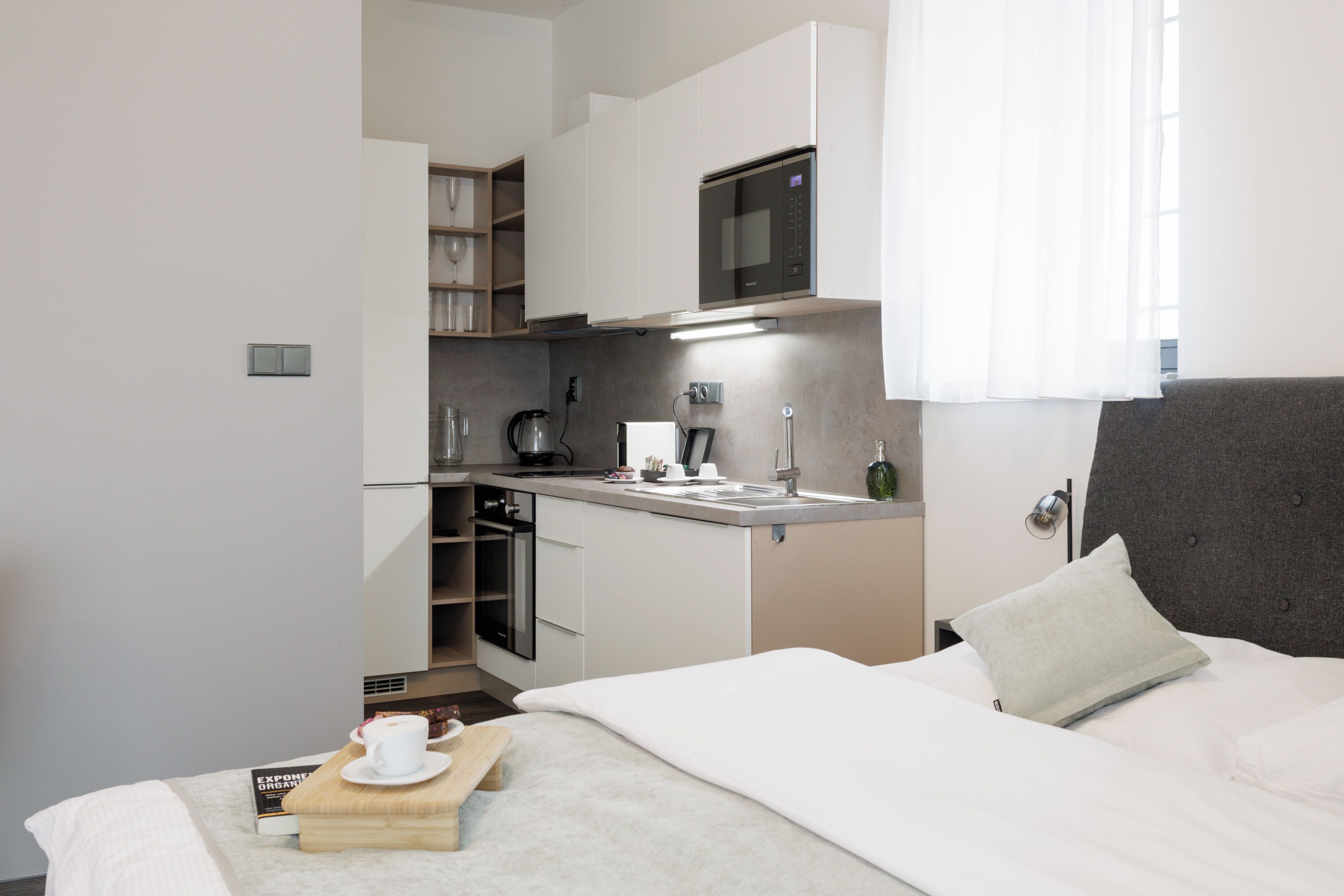 A compact hotel room with a kitchenette, a bed with a grey headboard, and a bedside table with a cup and saucer.