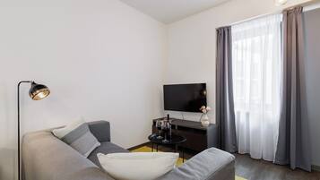 A modern living room with a grey sofa, a flat-screen TV, a small side table with wine glasses, and a floor lamp.