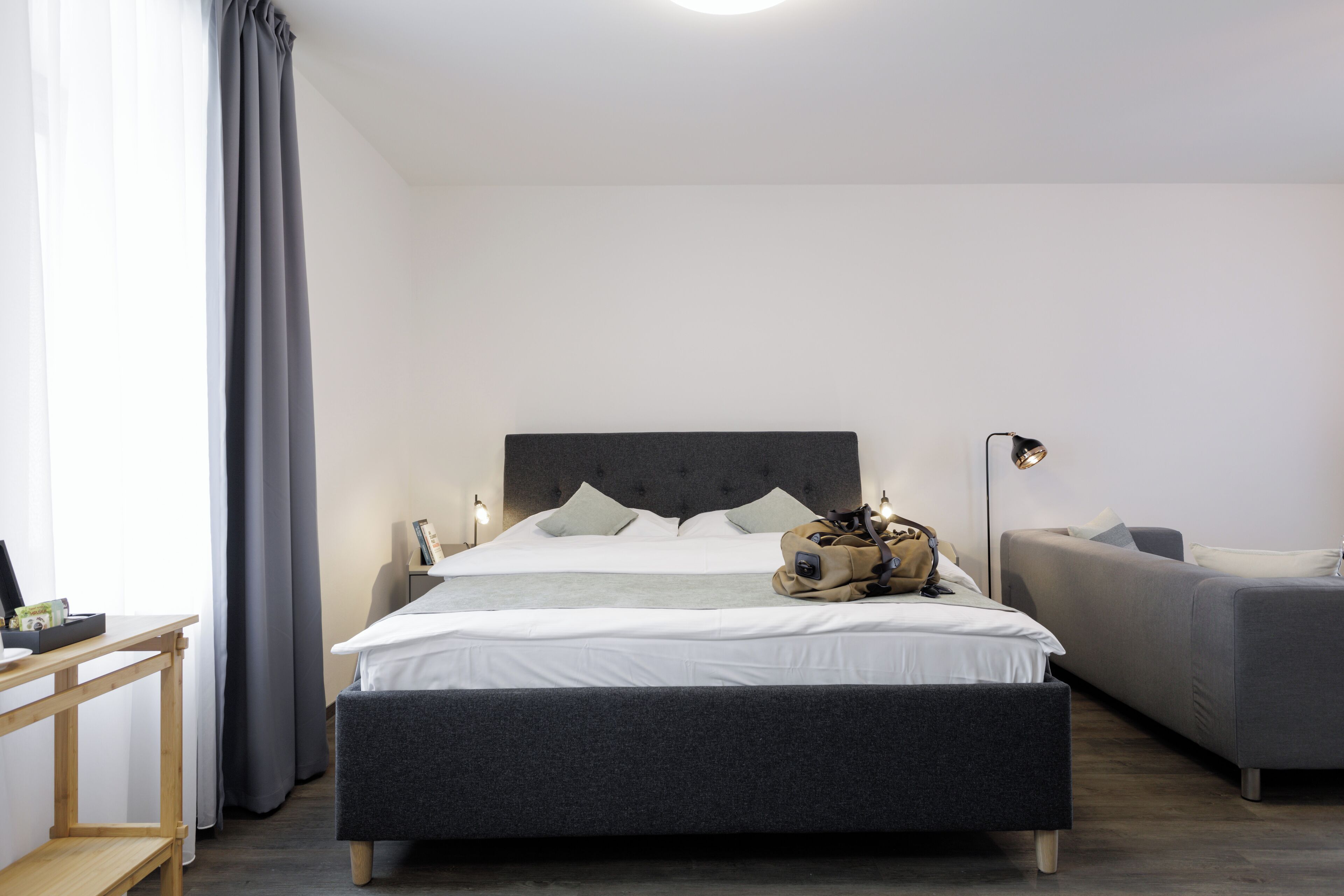 A modern bedroom with a large bed, a grey sofa, a wooden side table, and a floor lamp.