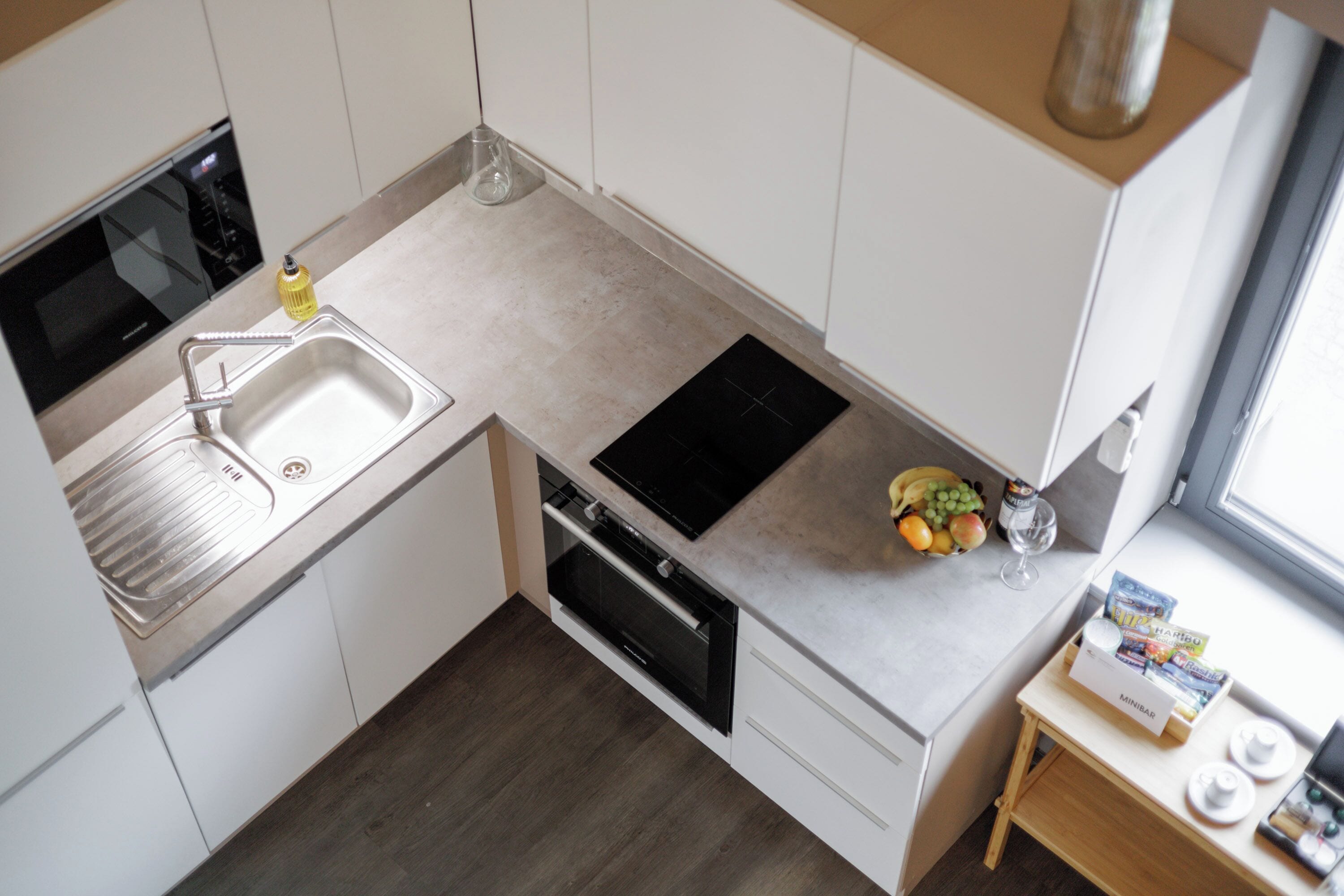 Una cocina moderna con microondas integrado, fregadero y horno. Hay una pequeña mesa de madera con un bol de fruta y una copa de vino.
