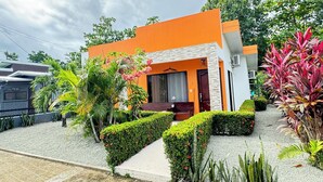 A modern building with a bright orange facade, a small porch, and a well-maintained garden with various plants and shrubs. - Süßes, Privates Bungalow mit Garden Nahe Manuel Antonio in Einer Ruhigen Gegend (San Martin Quepos)