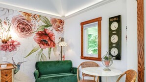 Een kamer met een groene bank, rieten stoelen, een ronde tafel, een bloemenwanddecoratie en een klok met daarop 