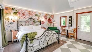 Een slaapkamer met bloemenbehang, een bed met metalen frame, een groene stoel, een kleine tafel en een klok aan de muur.