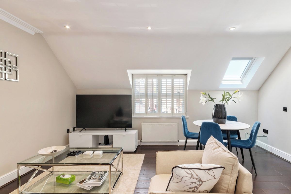 A modern living room with a glass coffee table, a sofa, a flat-screen TV, and a dining area with blue chairs and a white table.