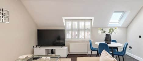 A modern living room with a glass coffee table, a sofa, a flat-screen TV, and a dining area with blue chairs and a white table.