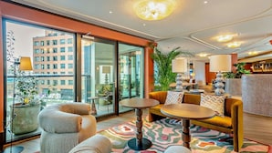 A modern hotel lobby with a large glass door leading to a balcony, a seating area with sofas and tables, and a decorative lamp. - Riverstone Heights (London)