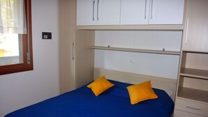 Quarto com uma cama, duas almofadas amarelas, uma janela com persianas e um guarda-roupa com vários compartimentos.