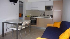 Uma cozinha compacta com micro-ondas, fogão e armários, uma mesa de jantar com cadeiras e um sofá com almofadas amarelas.