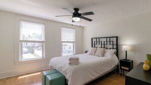 Une chambre à coucher avec un lit, un ventilateur de plafond, une table de chevet, une lampe et une fenêtre.