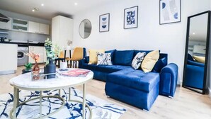 A modern living room with a blue sofa, a round coffee table, and a kitchen in the background. - 1 Bed Canary Wharf Flat (Greater London)