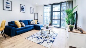 A modern living room with a blue sectional sofa, a round coffee table, and a large window offering a city view. - 1 Bed Canary Wharf Flat (Greater London)
