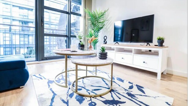 A modern living room with a large window, a blue sofa, a round coffee table, a TV on a white cabinet, and decorative plants. - 1 Bed Canary Wharf Flat (Greater London)