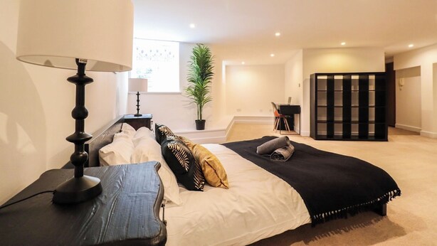 A bedroom with a bed, bedside table, lamp, potted plant, and a view of the outside. - Huge London 2 Bed Flat w/Parking (Greater London)