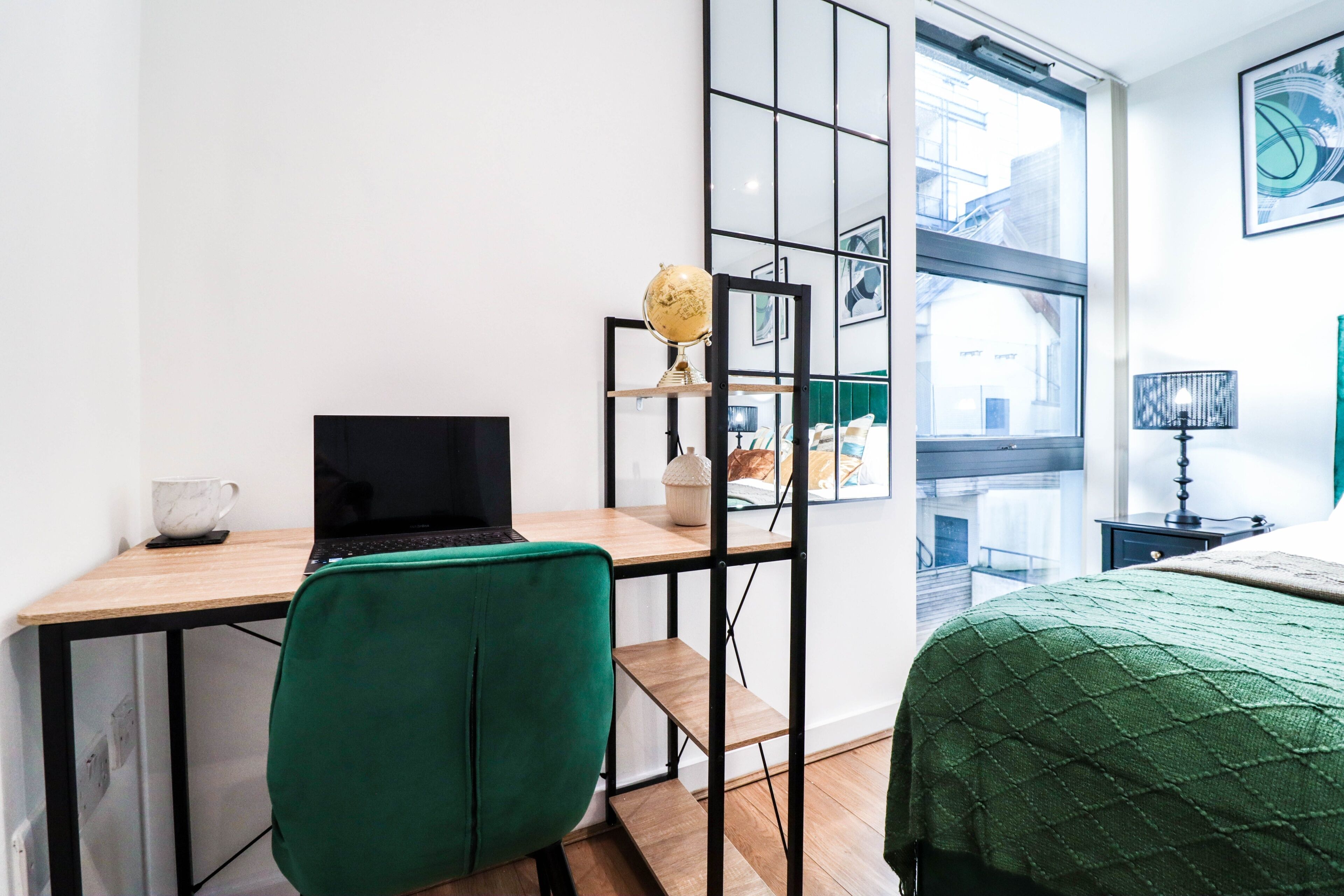 Una habitación moderna con un escritorio, una silla verde, una cama con colcha verde, un globo terráqueo en una estantería y una ventana con vista a la ciudad.