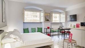A bedroom with a bed, a bedside table, a chair, a small table, a window, and a view of the outside. - Giustiniani Loft by Wonderful Italy (Genoa)
