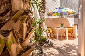 Eine Terrasse im Freien mit Tisch und Stühlen, umgeben von Pflanzen und einem bunten Sonnenschirm.