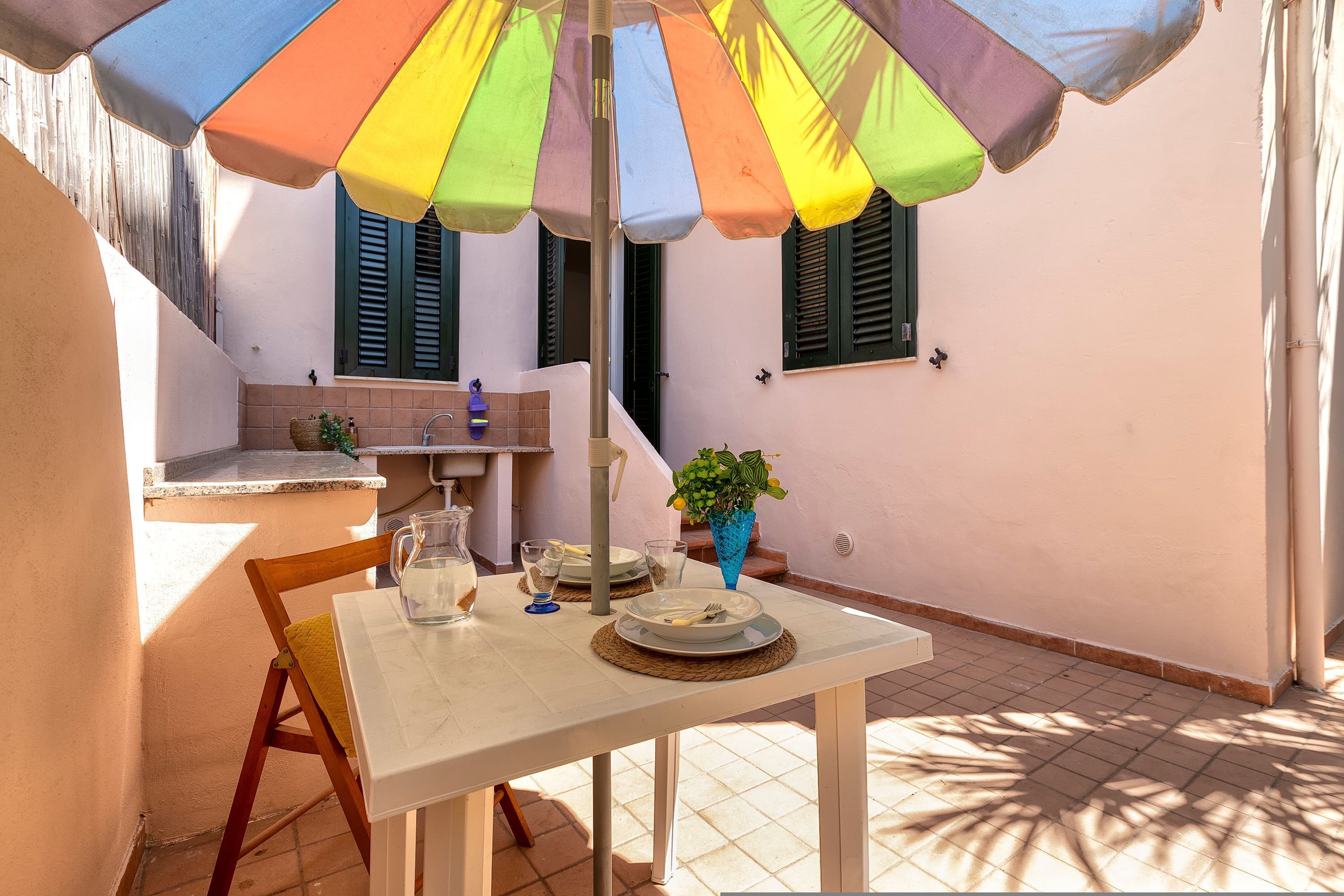 Une table à manger dressée avec des assiettes, des couverts et un pichet, placée sous un parasol coloré, avec une chaise et un pot de fleurs à proximité.
