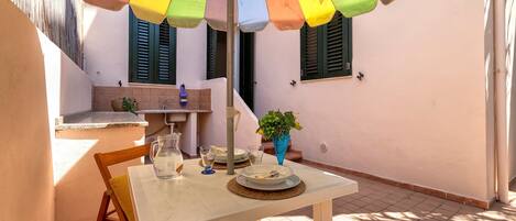 Une table à manger dressée avec des assiettes, des couverts et un pichet, placée sous un parasol coloré, avec une chaise et un pot de fleurs à proximité.