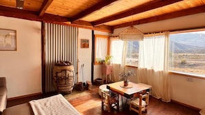 Ein gemütliches Zimmer mit Holzböden und hoher Decke, ausgestattet mit einer Couch, einem Esstisch mit Stühlen und Blick ins Freie.