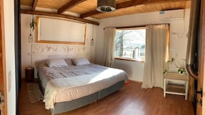 Ein Schlafzimmer mit Holzboden, einem Bett, einem Fenster mit Vorhängen, einem kleinen Tisch mit Pflanze und einer Heizung.