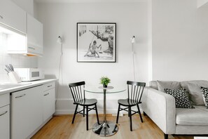 A modern kitchen with white cabinets, a microwave, and a round glass table with two chairs. A framed black and white photo hangs on the wall above the table. - Marylebone by Excel Apartments (London)