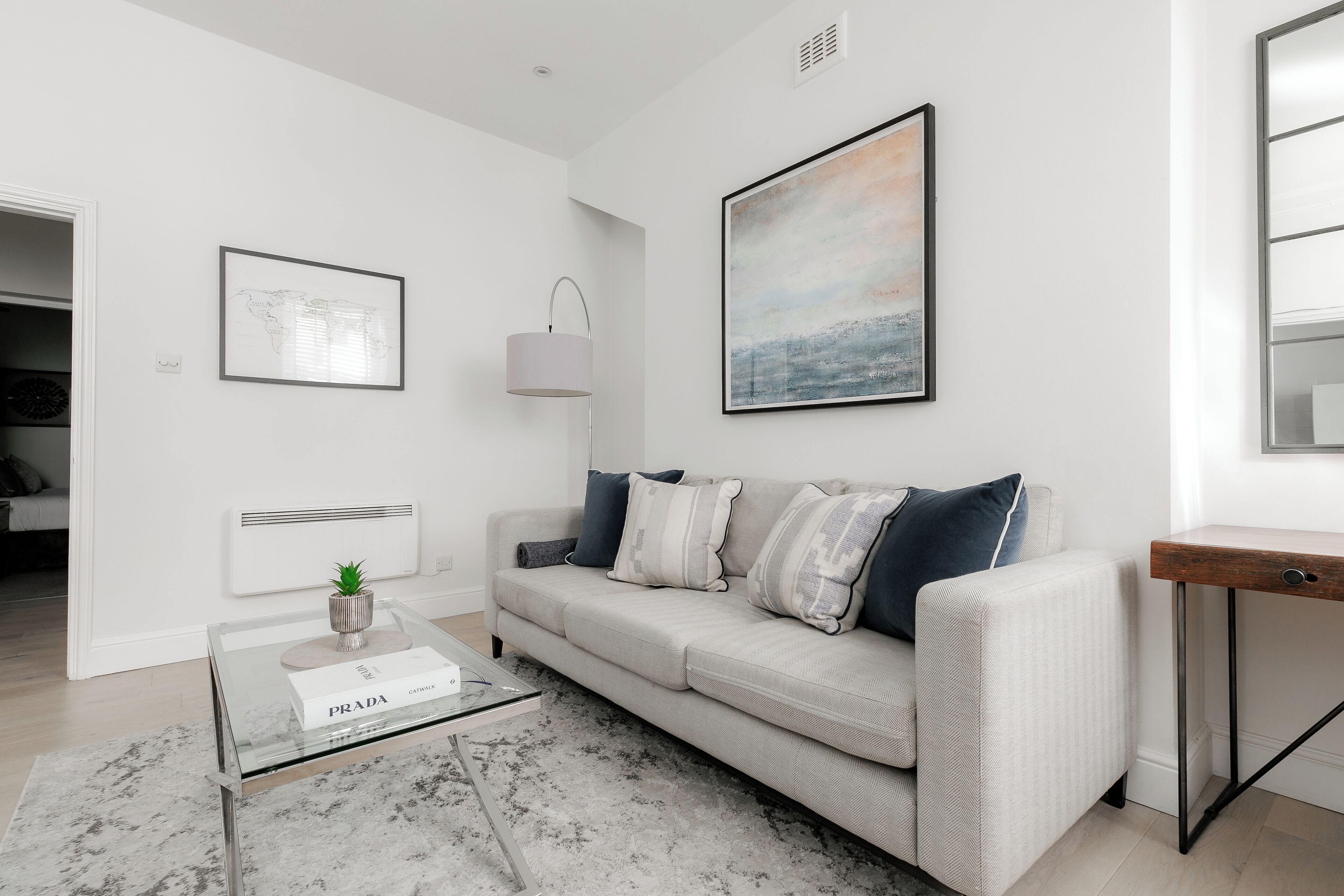 A modern living room with a light-colored sofa, a glass coffee table with a Prada item, a world map, a landscape painting, and a floor lamp.