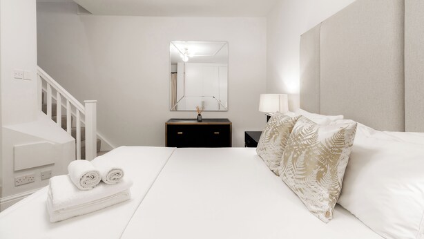 A hotel room with a neatly made bed, a bedside table with a lamp, a mirror above the dresser, and a staircase to the left. - Marylebone by Excel Apartments (London)