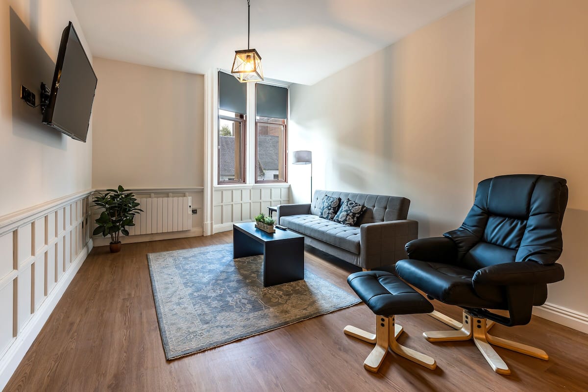 A modern living room with a flat-screen TV mounted on the wall, a black leather chair, a grey sofa, a wooden coffee table, and a patterned rug.