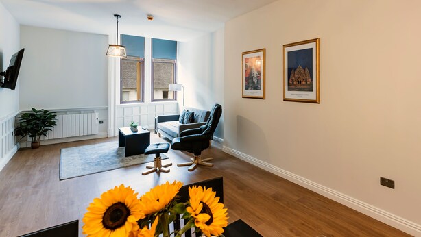 A modern living room with a sofa, armchairs, a coffee table, and a dining table with sunflowers. - The Hazlitt Apartments (Paisley)